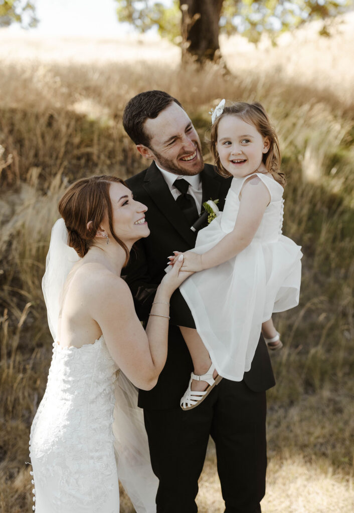 Wedding couple holding little girl while all smile with golden field behind them at French Oak Ranch