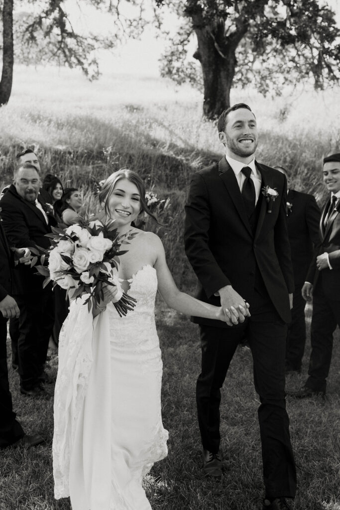 Wedding couple holding hands while smiling and walking through groomsmen and bridesmaids towards camera at French Oak Ranch