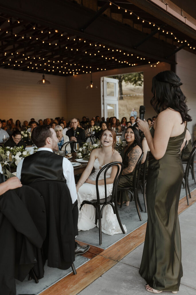 Bridesmaid giving speech during wedding reception while couple looks at her and bride laughs at French Oak Ranch