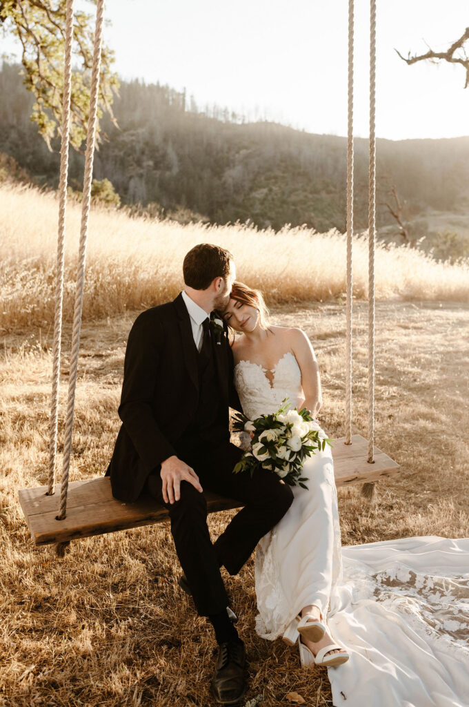 Wedding couple sitting on wooden rope swing while groom kisses bride on head with golden field behind them at French Oak Ranch