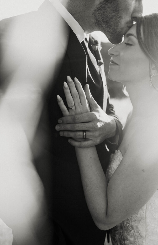 Close up of groom kissing bride on forehead while holding her hand on his chest with wedding rings at French Oak Ranch