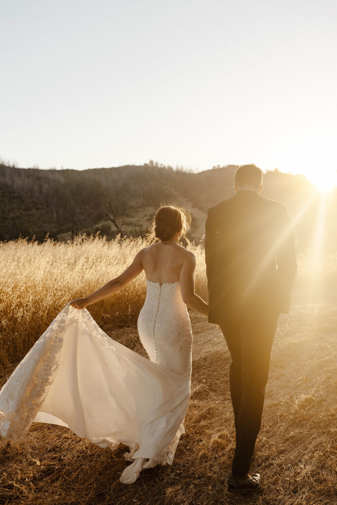 Wedding couple holding hands while bride plays with dress tail and walking along dirt path with sun in background at French Oak Ranch