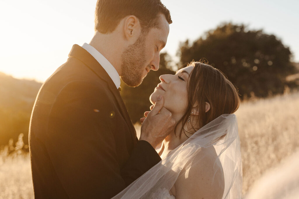 Groom holding brides face while both smile with their eyes closed and golden field behind them at French Oak Ranch