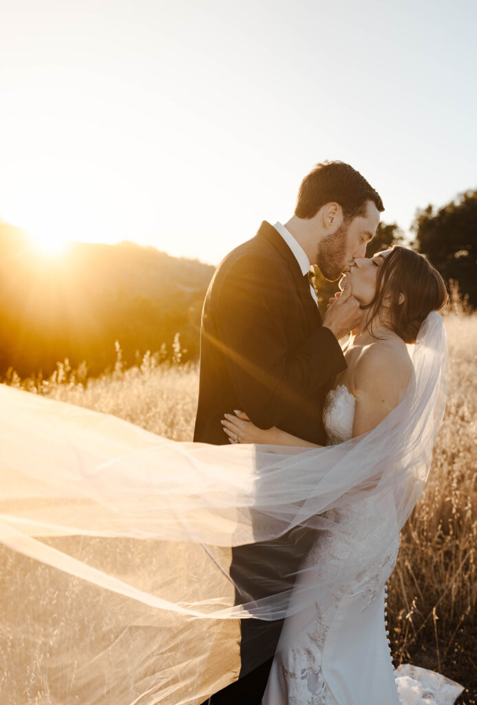 Groom holding wife's chin as wedding couple kisses in golden field with sun behind them at French Oak Ranch 