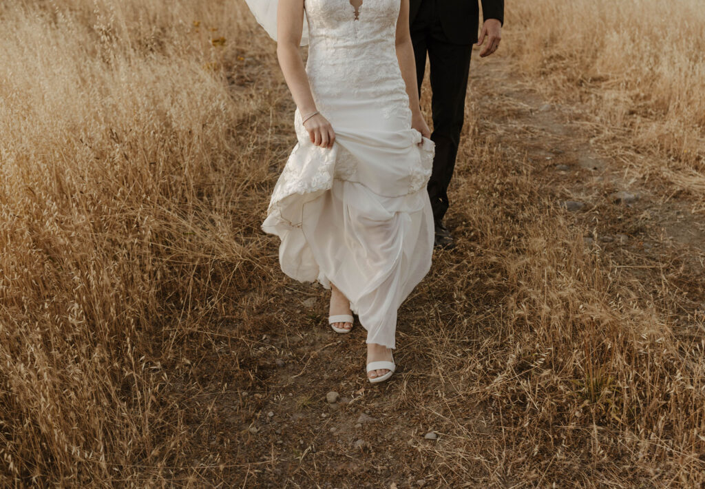 Bride holding dress as wedding couple walks through dried grass at French Oak Ranch