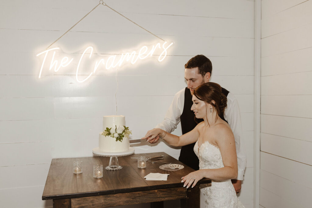 Wedding couple holding knife together while cutting wedding cake on wooden table with neon wedding sign with last name hanging above them at French Oak Ranch