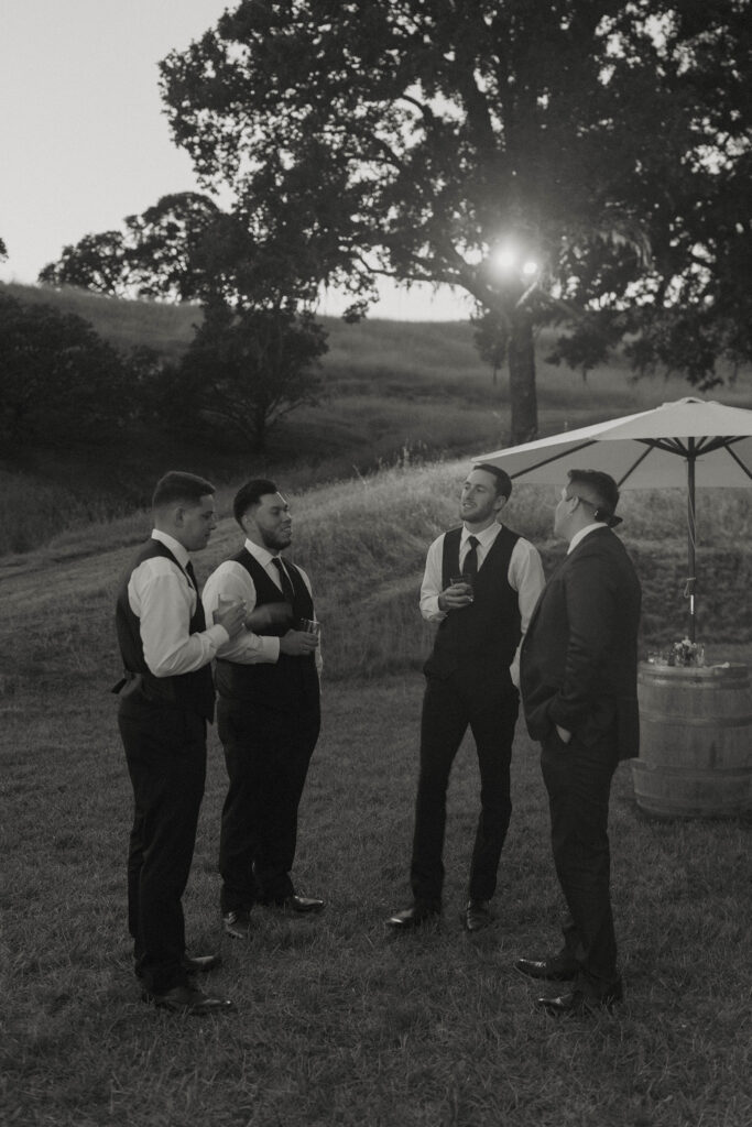 Wedding groom standing and talking with groomsmen while holding drink with golden field and sparse trees behind them at French Oak Ranch