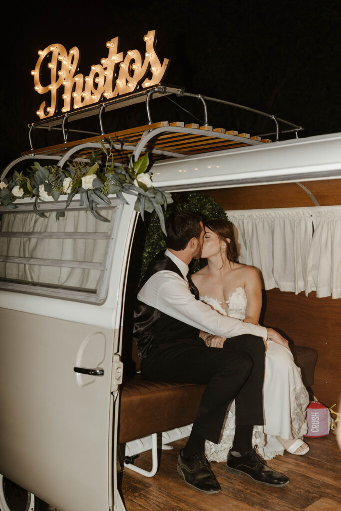 Wedding couple kissing while inside photobooth inside of white van with lighted "photos" sign on top at French Oak Ranch