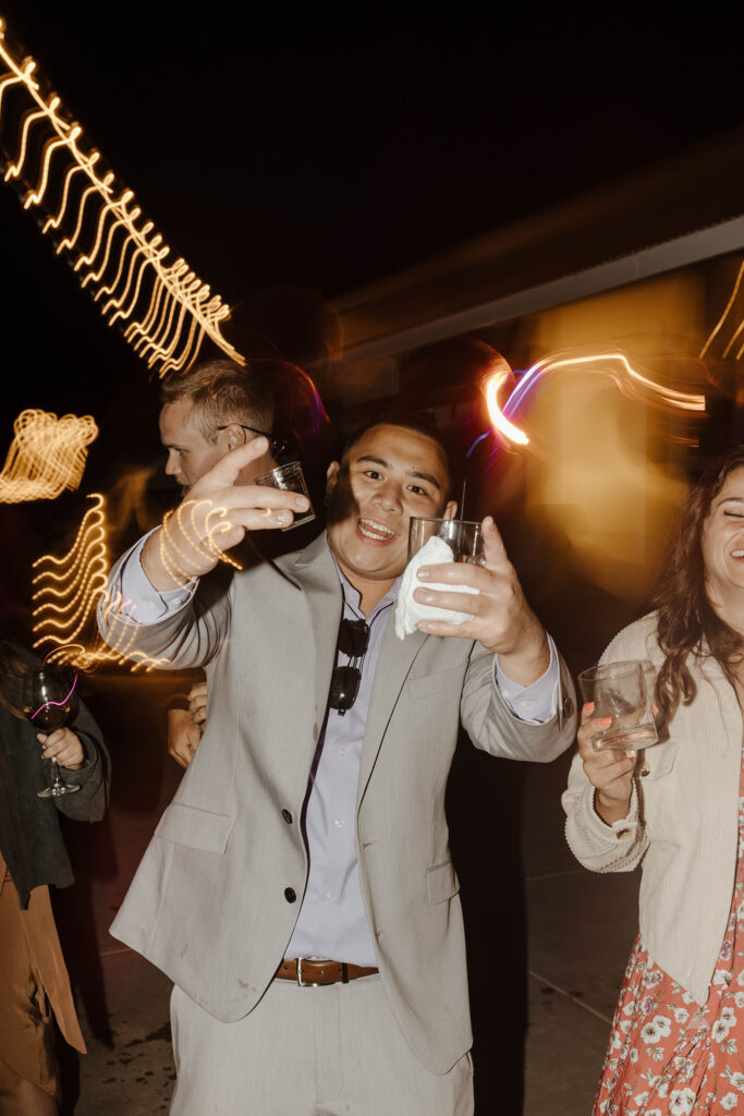 Wedding guests celebrating while holding drinks toward camera with blurred lights in background at French Oak Ranch