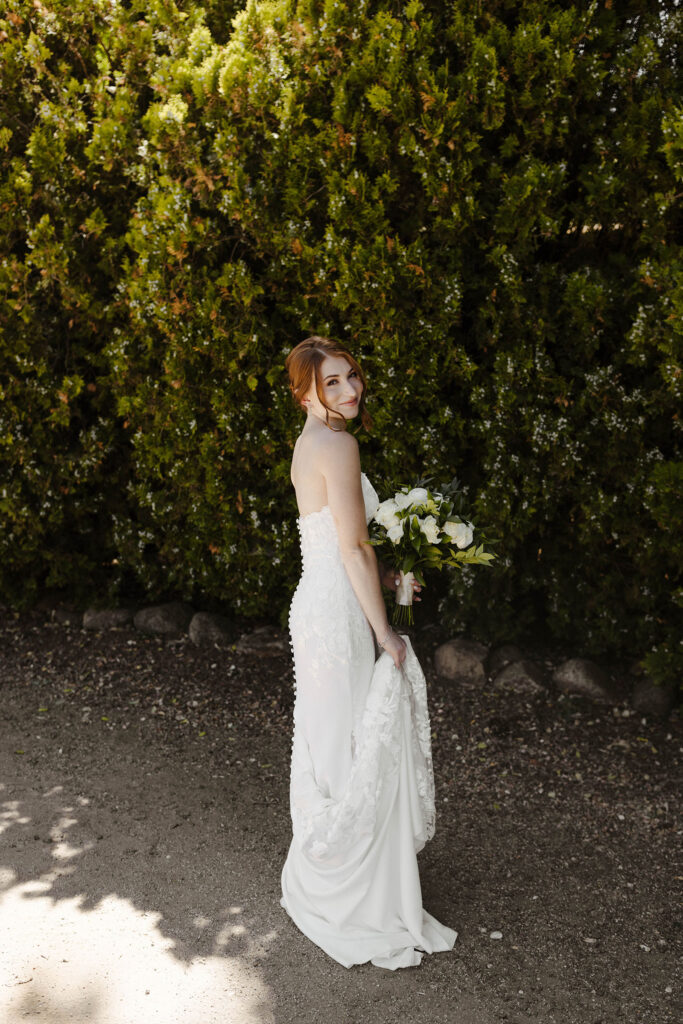 Wedding bride holding dress and smiling while looking back over shoulder with tall bushes behind her at French Oak Ranch