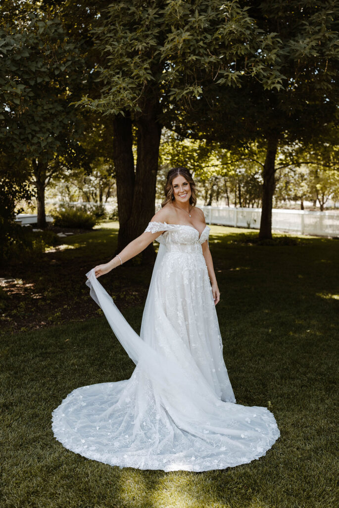 Bride holding trail of wedding dress while smiling at camera and standing in grass with trees in background at Jacobs Berry Farm