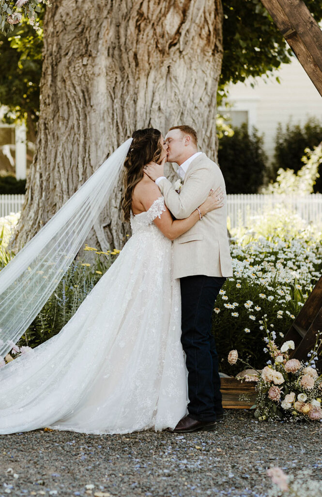 Wedding couple kissing during ceremony while groom holds bride face in front of large tree and white flowers at Jacobs Berry Farm