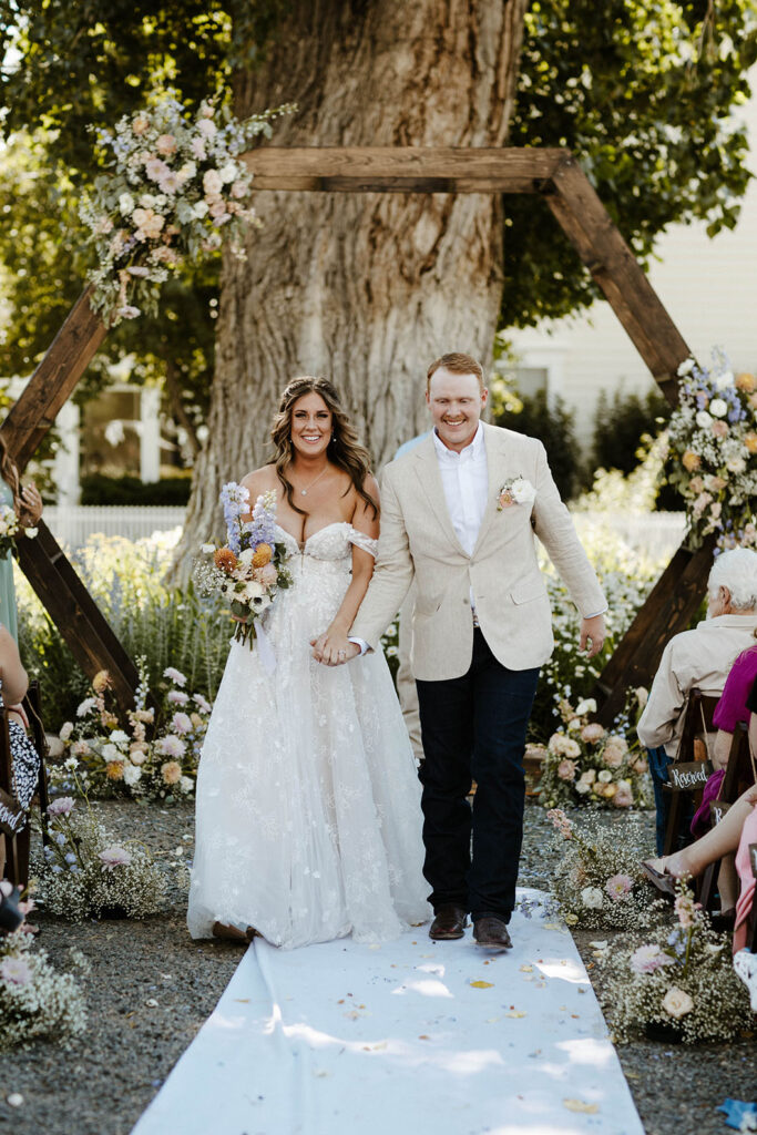 Wedding couple holding hands and smiling while walking down aisle together after ceremony with colorful florals behind them at Jacobs Berry Farm