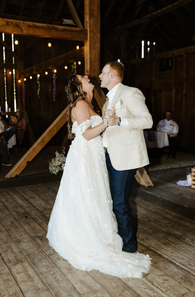 Wedding couple holding hands while dancing and bride is laughing inside wooden barn with lights at Jacobs Berry Farm