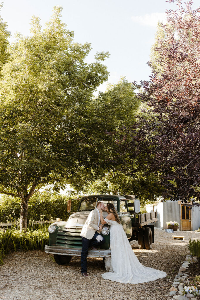 Wedding couple kissing in front of old green pickup truck with tall purple and green trees above them at Jacobs Berry Farm