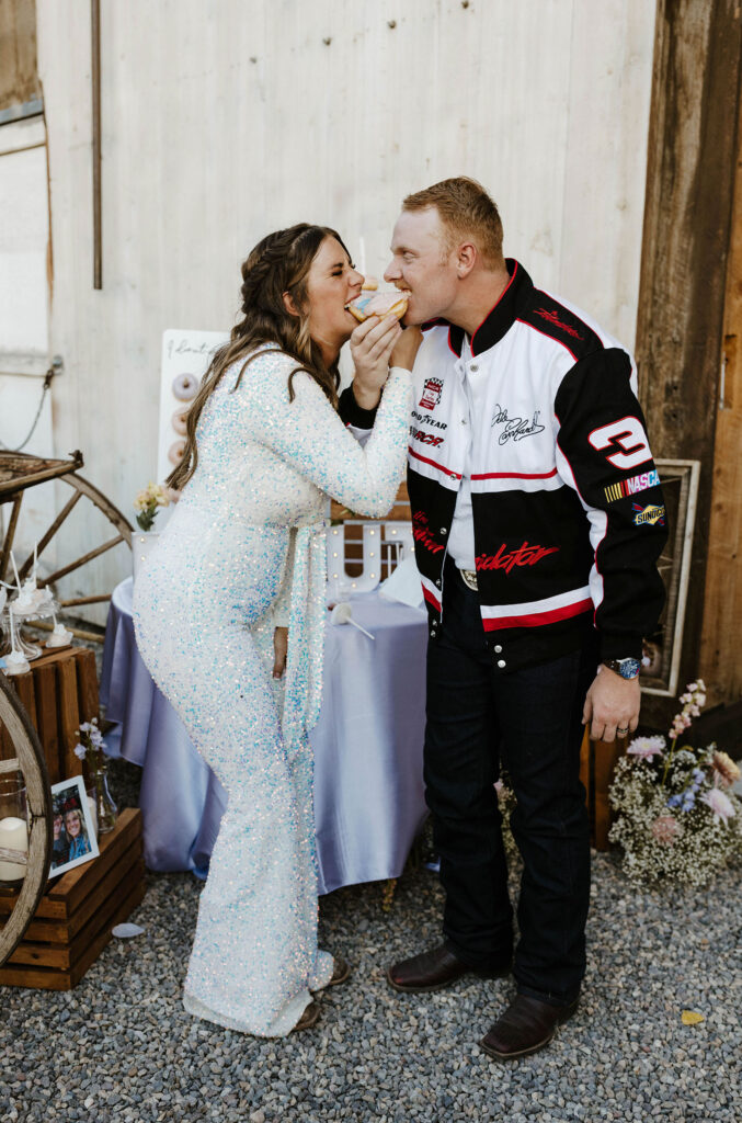 Wedding couple eating doughnuts with arms intertwined while bride is in reception outfit and groom wears nascar jacket at Jacobs Berry Farm