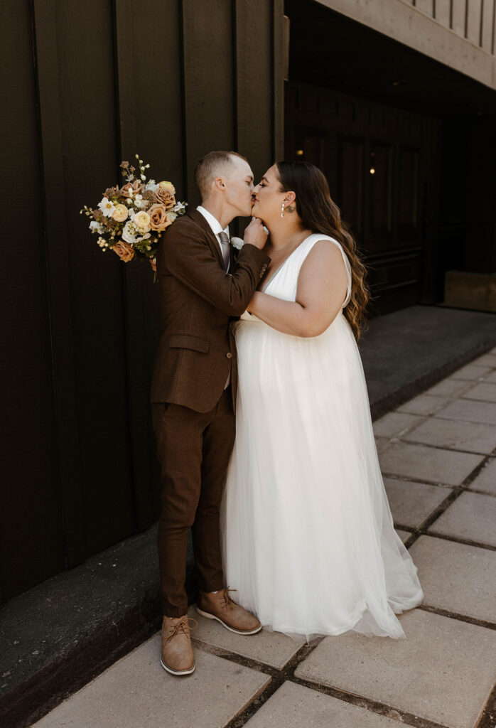 Wedding couple kissing while holding each other's faces at the coachman hotel with brown and pink floral bouquet