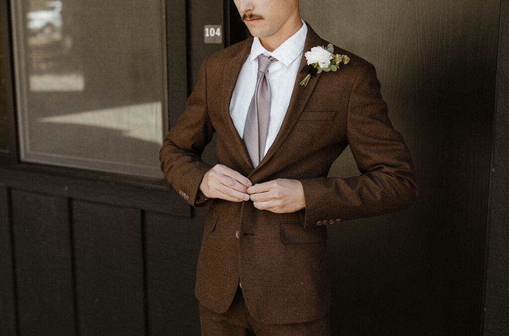 Wedding groom buttoning suit jacket while standing in front of dark brown building at the Coachman hotel in Lake Tahoe