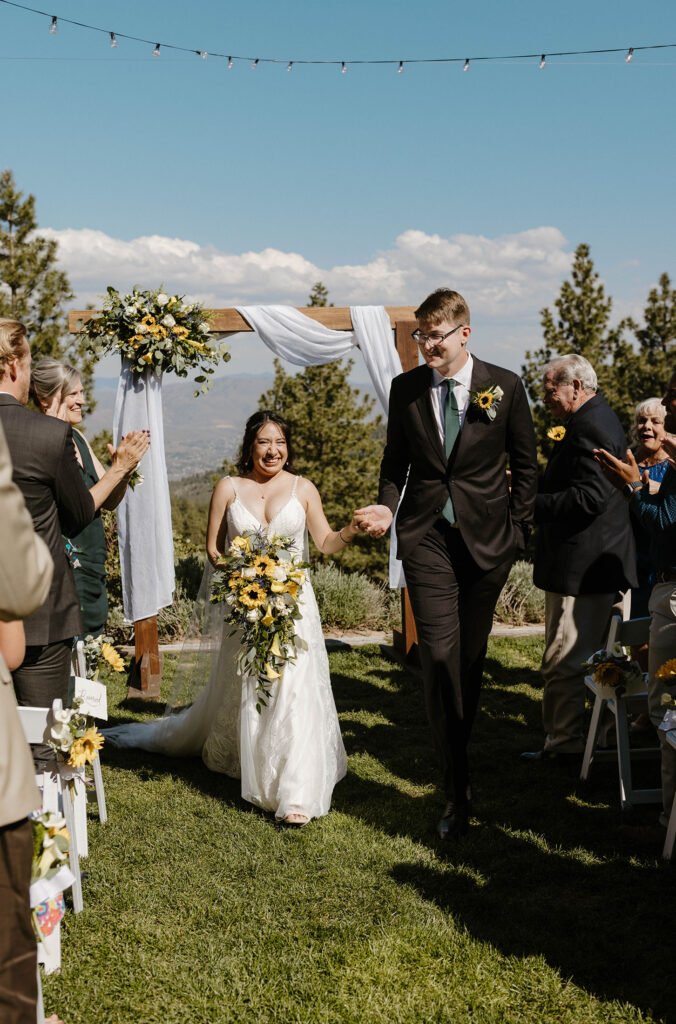Wedding couple holding hands while walking down aisle together after ceremony while bride holds sunflower bouquet at the Tannenbaum