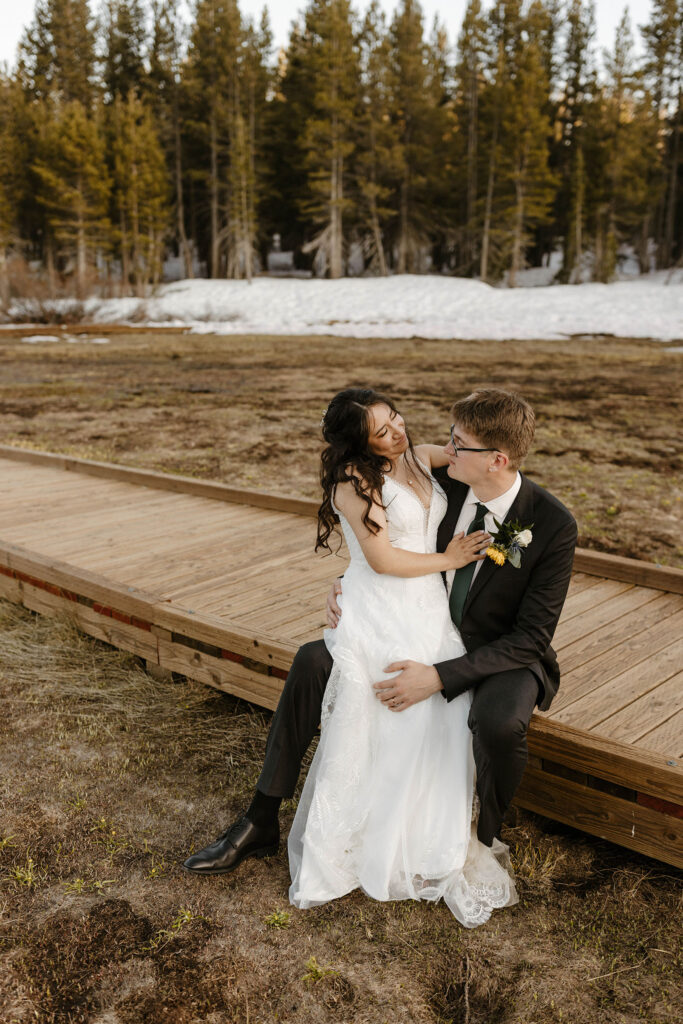 Wedding bride sitting on grooms knee as couple sits on wooden walkway with snow in background at the Tannenbaum