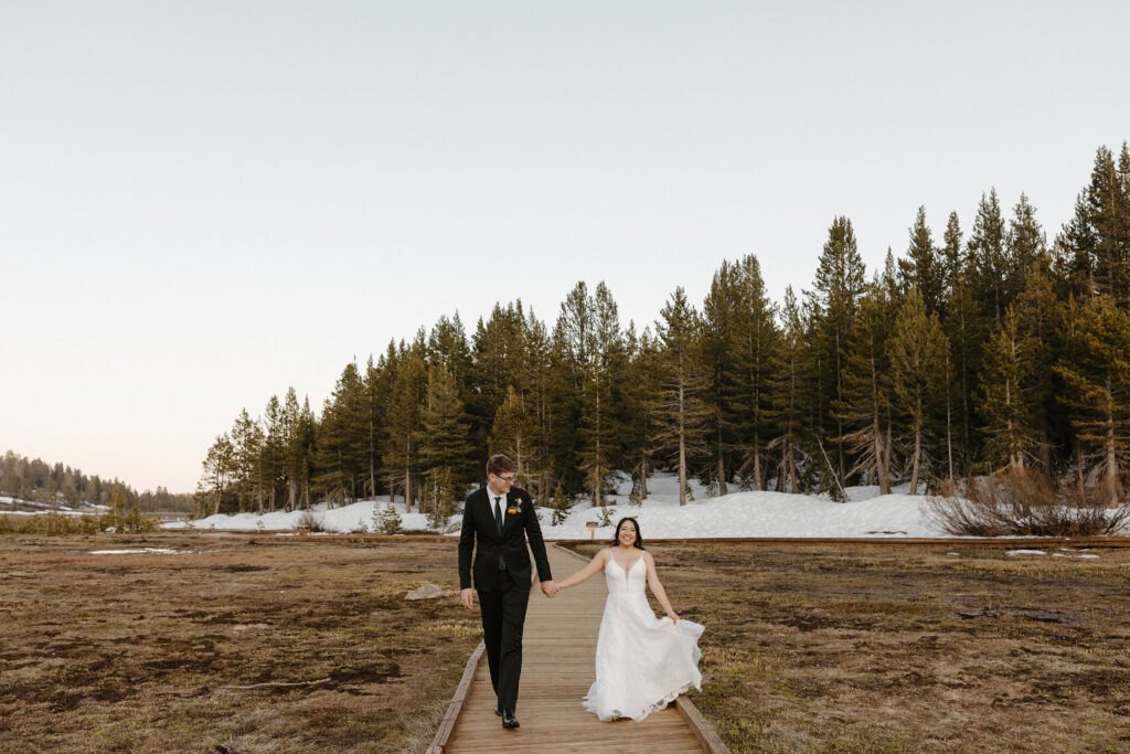 Wedding couple holding hands while walking along wooden walkway together towards camera with pine trees and snow in background at the Tannenbaum