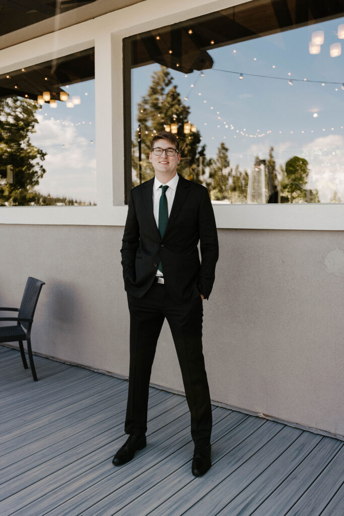 Wedding groom in wedding attire standing outside large window looking at camera at the Tannenbaum