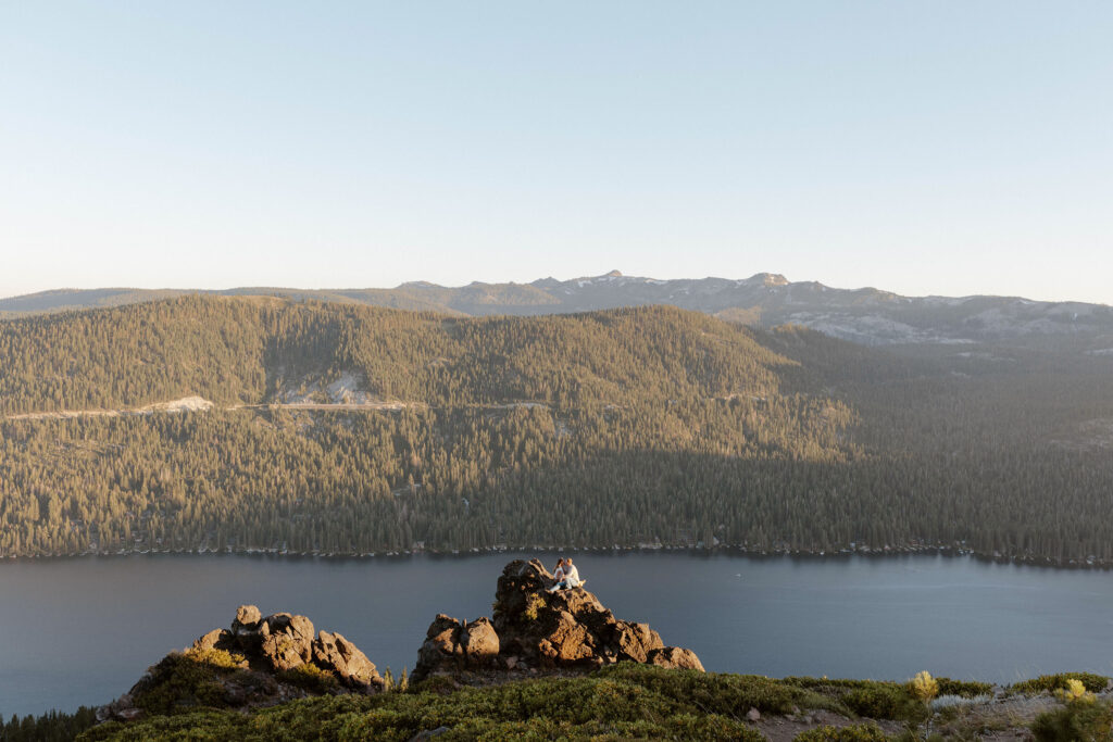 Far away shot of married couple sitting on large rock formation and holding each other with Donner Lake in background and below with pine trees