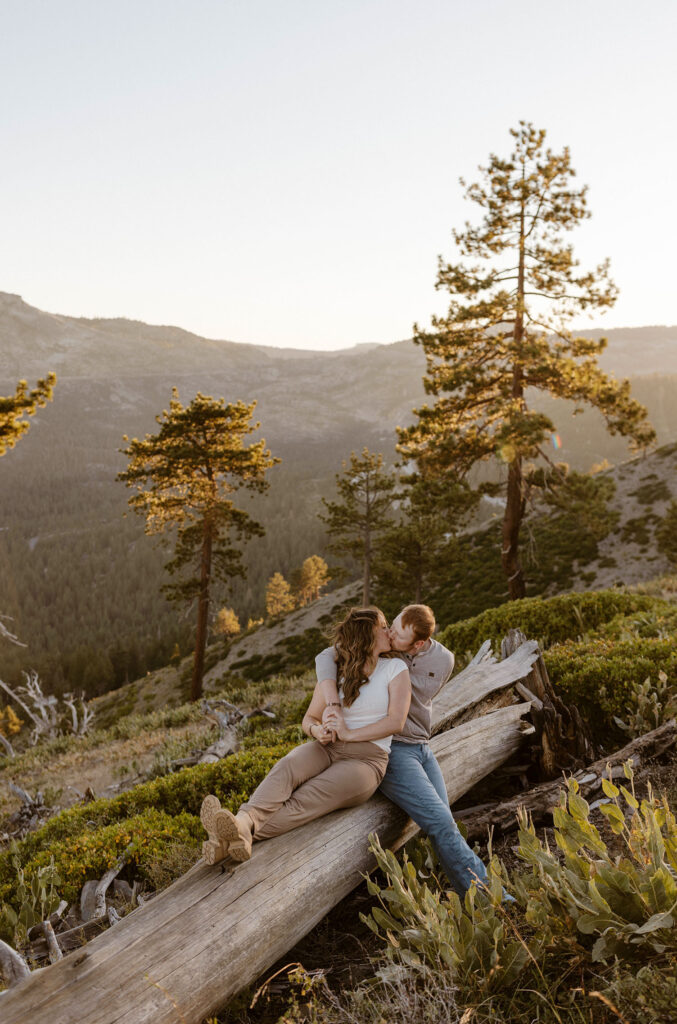 Married couple sitting on log together while holding hands and kissing with mountains and pine trees in background at Donner Lake