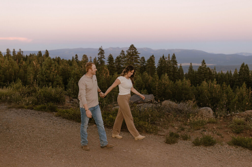 Married couple holding hands while walking down dirt trail together and smiling with purple mountains and pine trees in background at Donner Lake
