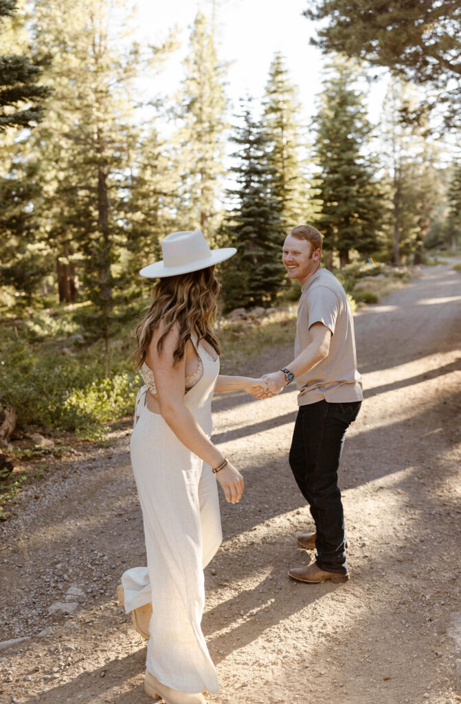 Married couple holding hands on dirt trail while man smiles at Donner lake with pine trees in background