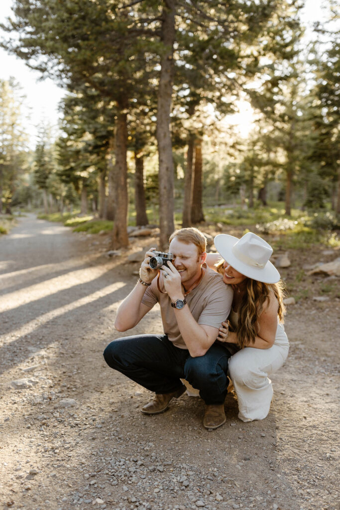 Man playing with camera while wife crouches behind him on dirt trail and both laugh at Donner Lake