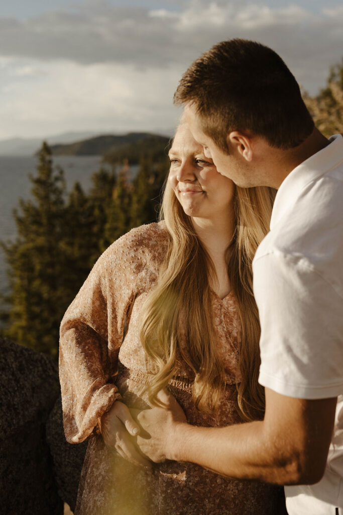 man holding woman's stomach and kissing her forehead while she looks at lake tahoe