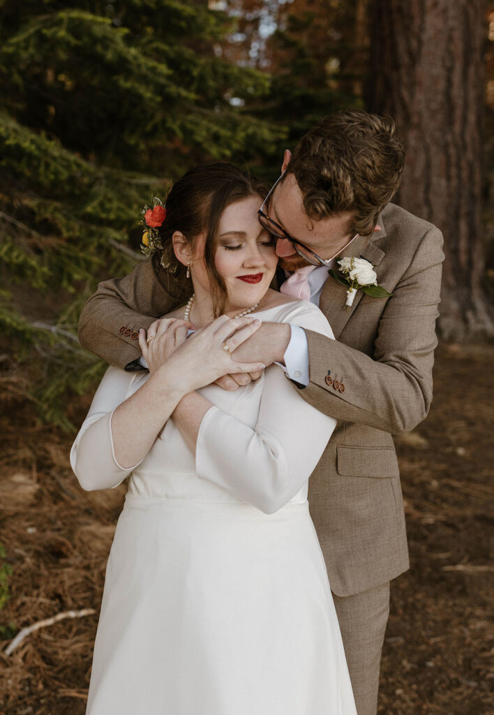 Groom hugging bride from behind while she holds his hands and smiles at Valhalla in Lake Tahoe