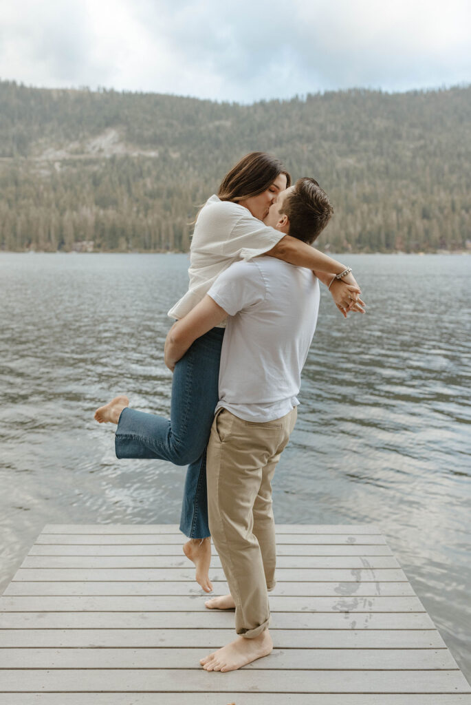 Man lifting fiancé up while both kiss on a pier in lake Tahoe with pine trees and lake in background