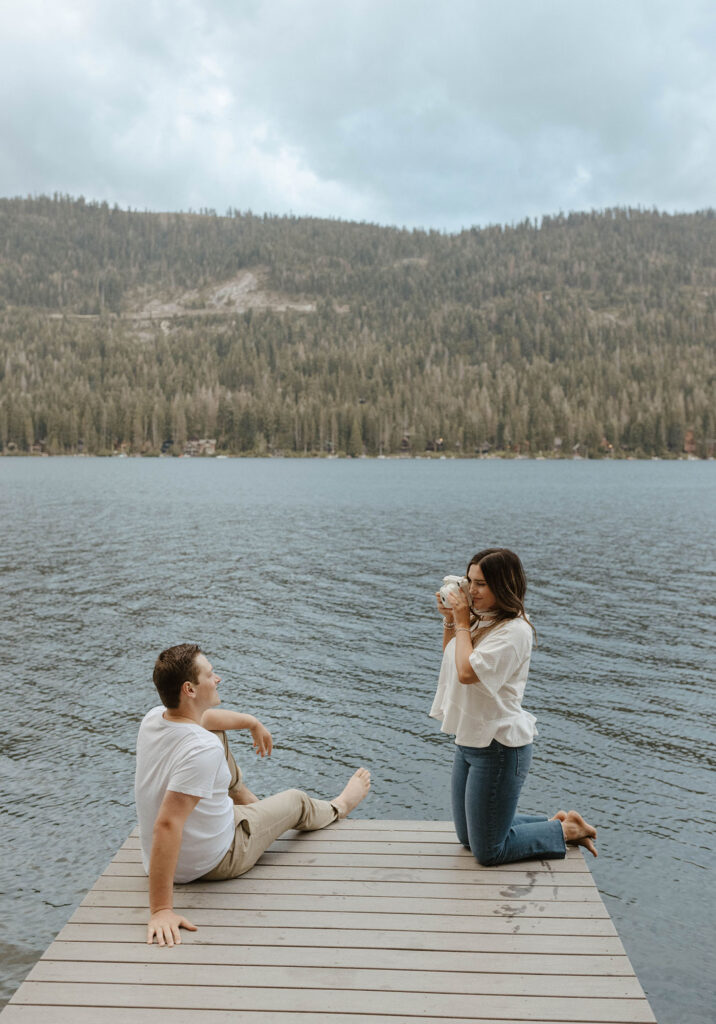 Woman taking picture of fiancé while they both are sitting on a pier in Lake Tahoe with pine trees in background