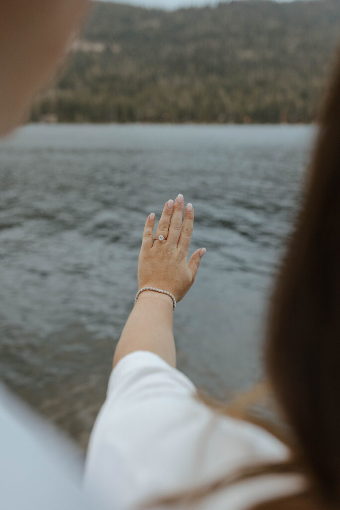 Woman admiring engagement ring on hand in Lake Tahoe with pine trees and lake in background