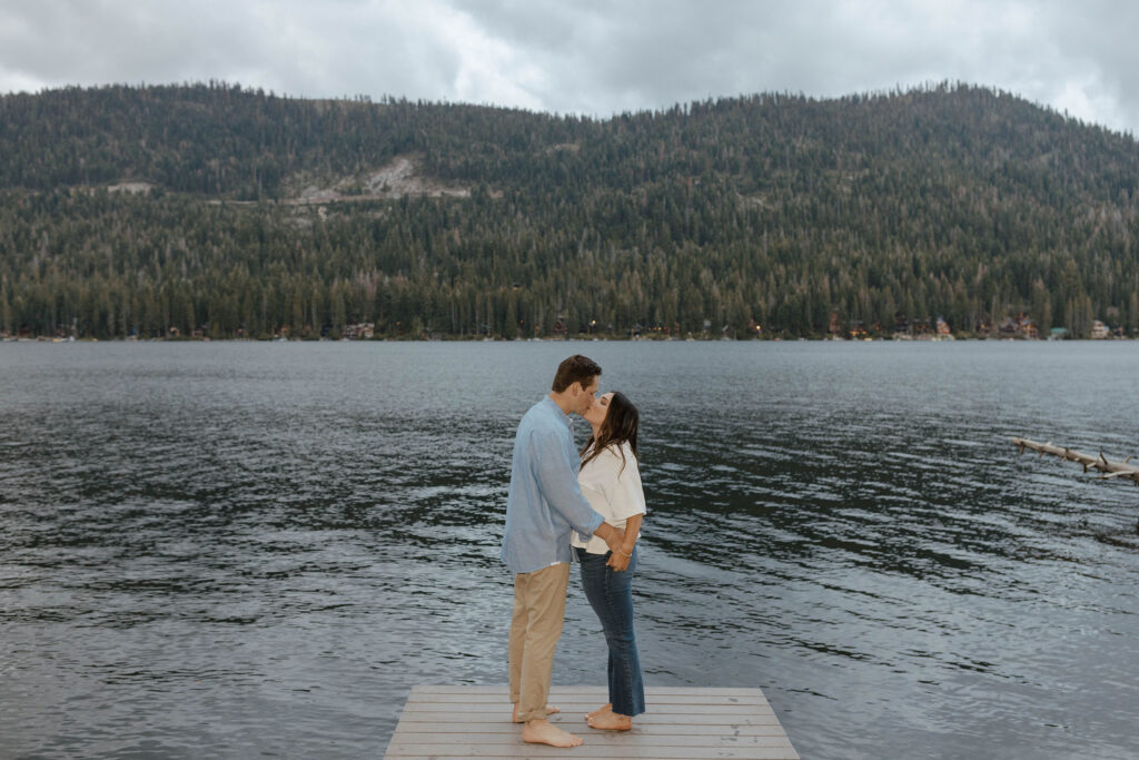 Engagement couple holding each other and kissing while standing on pier in Lake Tahoe with pine trees and mountains in background