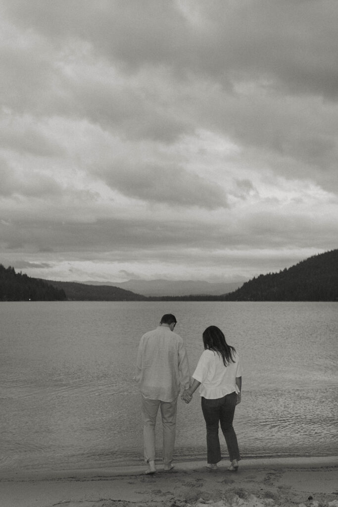 Engagement couple holding hands on sandy beach walking towards water together in Lake Tahoe