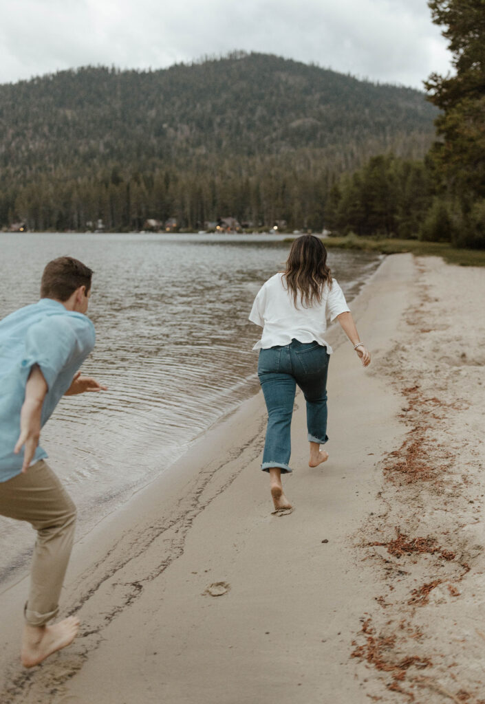 Man chasing fiancé down sandy beach in Lake Tahoe with pine trees and mountains in background