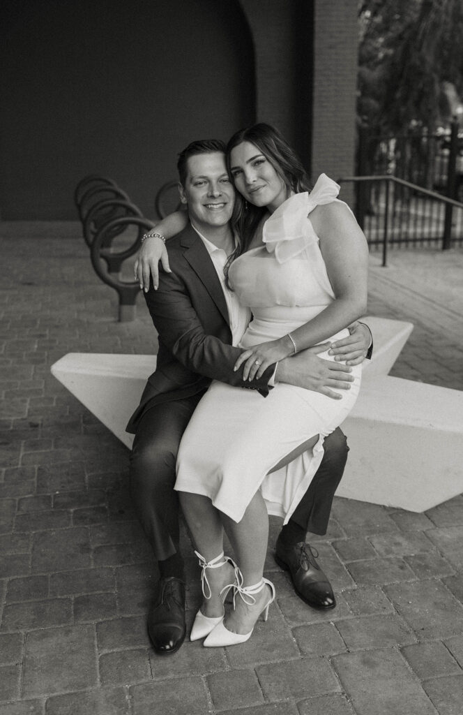 Engagement couple sitting on concrete bench together while smiling at camera in downtown Reno