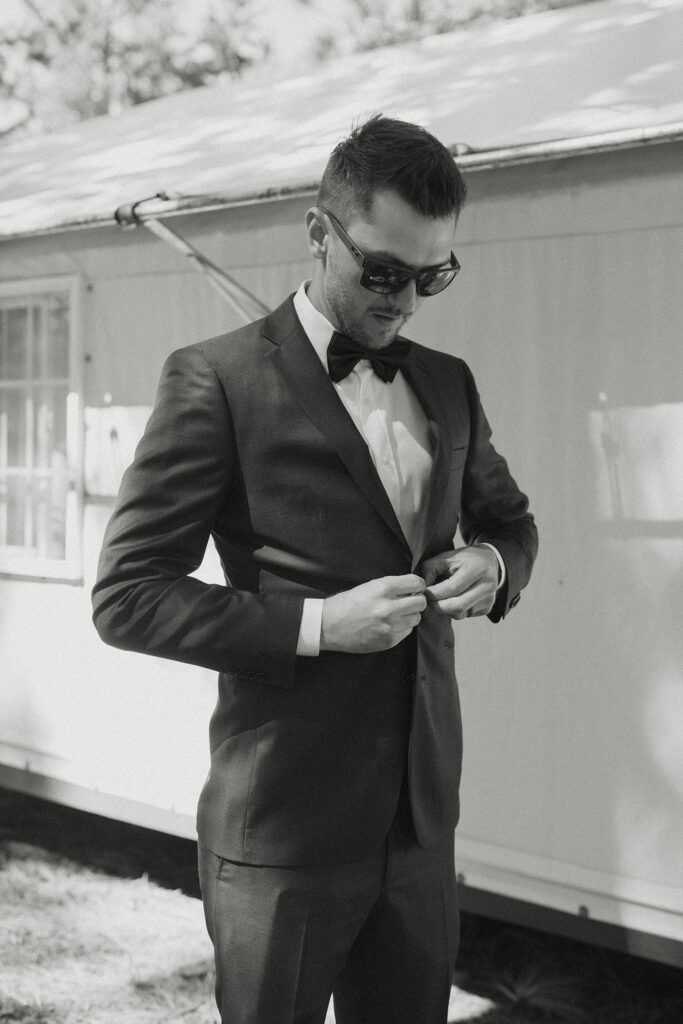 Wedding groom buttoning up jacket while wearing sunglasses outside of canvas cabin at dancing pines