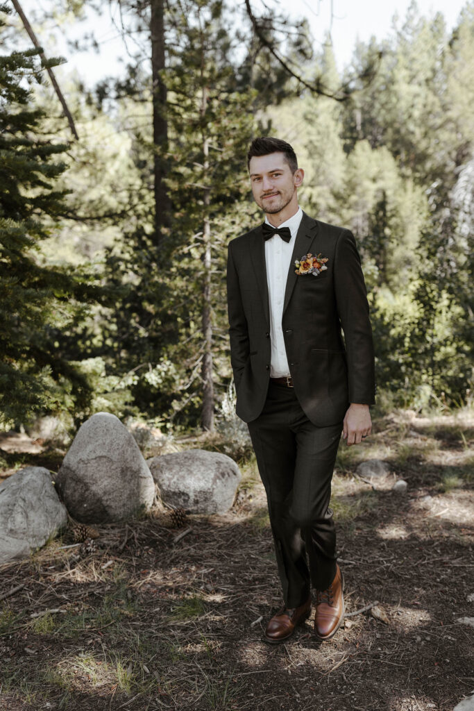 Wedding groom smiling at camera while walking along trail with hand in pocket in front of pine trees at dancing pines