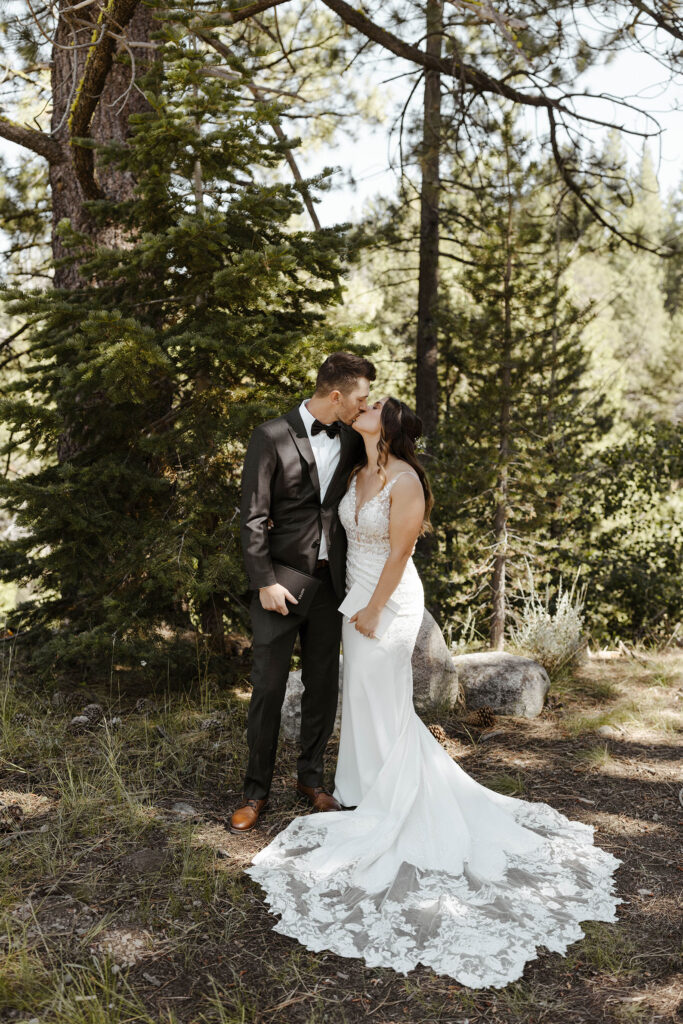 Wedding couple holding vow books while kissing in front of pine trees at dancing pines