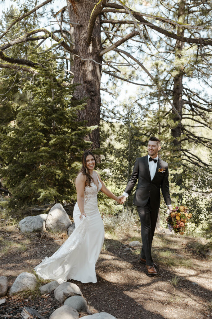 Wedding couple holding hands while walking along trail together and smiling surrounded by pine trees at dancing pines