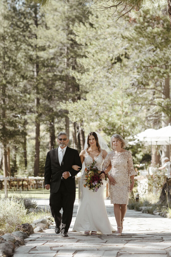 Wedding bride walking down aisle while holding arms with mom and dad at dancing pines