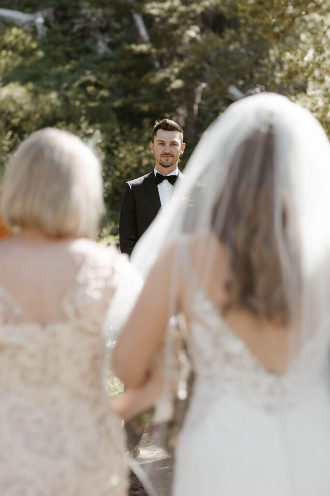 Groom watching bride walk down wedding aisle while holding arms with parents at dancing pines