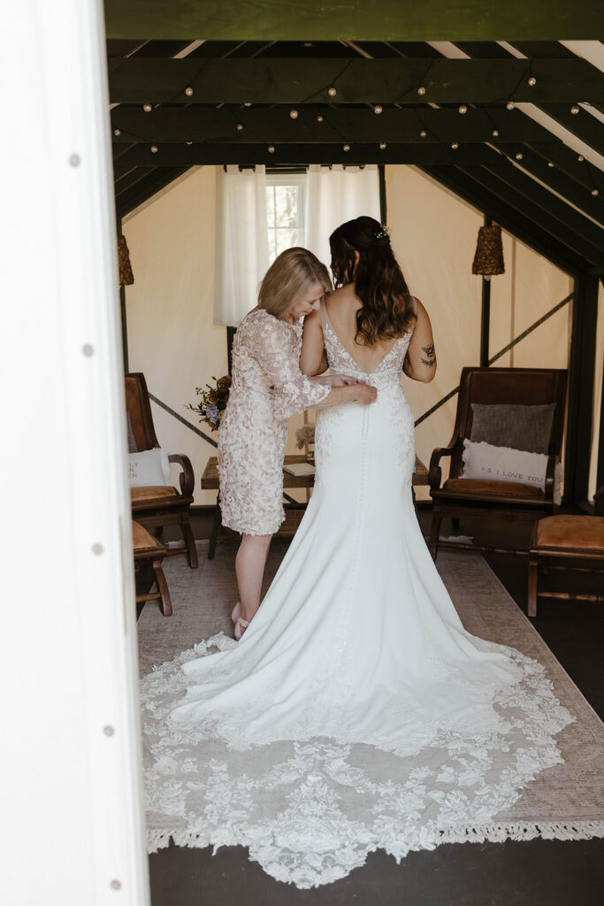 Mom helping bride button up back of wedding dress while inside at dancing pines