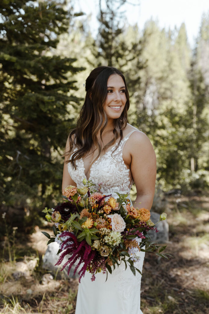 Wedding bride holding multicolored floral bouquet while smiling in front of pine trees at dancing pines