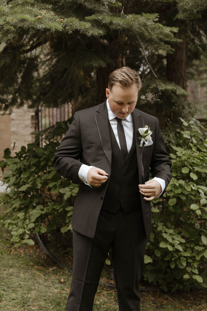 Wedding groom looking down and buttoning suit jacket while in front of tree and greenery at Aspen Grove