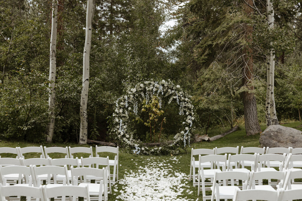 Wedding arch with hanging white flowers in front of white chairs and flower petals at Aspen Grove with tall trees behind